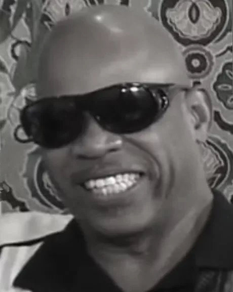Bold black man with a dark sunglasses and a big toothy smile.