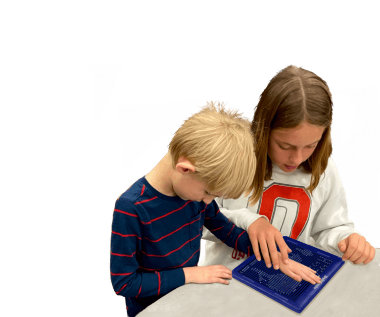 A boy with blonde hair about seven years old and wearing a blue stripe shirt, is standing with his big sister about 11 and shoulder length, with straight brown hair, in a white sweatshirt is guiding his hand on the BrailleDoodle to feel the Braille.