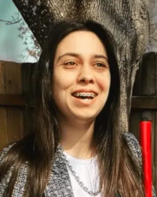 Woman in her 20s with long dark hair, wearing a blazer and a white blouse, smiling outdoors and holding her white cane