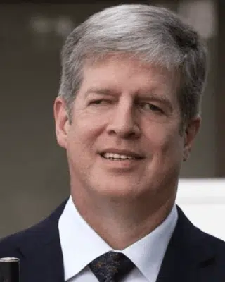 Middle-aged man smiling with grey hair, in a dark suit is holding a white cane